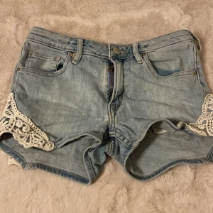 Ljusblå jeansshorts med spets från H&M - Snygga ljusblå jeansshorts från H&M med coola spetsdetaljer i vitt på sidorna. Klassisk femficksmodell, bälteshällor och dragkedja framtill. Perfekta för varma dagar när du vill ha en chill och trendig look.