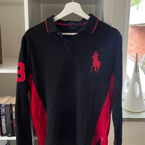 Svart långärmad pikétröja Polo Ralph Lauren - Svart långärmad pikétröja från Polo Ralph Lauren med röd broderad logga på bröstet och röd siffra 3 på ärmen. Röd detalj på kragen och sidorna. Klassisk krage med knappar och sportig vibe. Perfekt för dig som gillar stilrena streetlooks och golf. Finns en vit liten fläck längst ner på tröjan.