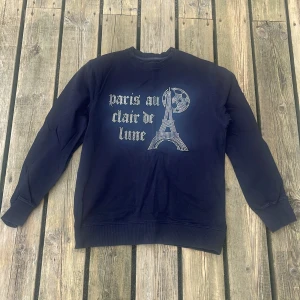 Mörkblå sweatshirt med strassmotiv - Mörkblå sweatshirt från BLINGLUCID i bomull med lång ärm. Framsidan har ett glittrigt strassmotiv med texten 'paris au clair de lune' och Eiffeltornet under en måne. Klassisk passform och ribbade muddar vid ärmslut och nederkant.