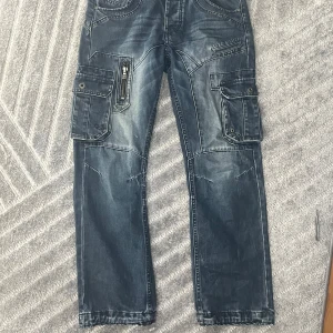 ETO blå cargopants med detaljer - ‼️FRÅGA OM BYXORNA FINNS INNAN KÖP‼️ Säljer ett par blå cargopants från ETO med coola kontrastsömmar, flera stora fickor med lock och dragkedja samt broderad logga bak. Jeansen har en loose fit och är tillverkade i denim med slitna detaljer för en streetig look.