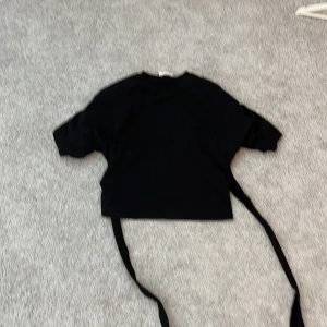 Svart croppad topp med knyt från Nelly - Svart croppad topp från Nelly med korta ärmar och långa knytband i ryggen. Toppen har en enkel och clean design med rund halsringning och passar perfekt till jeans eller kjol för en trendig look.