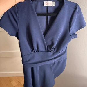 Mörkblå omlottklänning från Blume - Supersnygg mörkblå kortklänning från Blume London med omlott och v-ringning. Klänningen har korta ärmar och markerad midja som ger en smickrande siluett. Perfekt för dig som gillar stilrena och trendiga plagg.