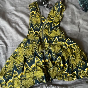 Gul och svart mönstrad kortklänning - Säljer en färgstark kortklänning med coolt grafiskt mönster i gult, svart och vitt. Klänningen är ärmlös med breda axelband och har en luftig passform. Perfekt för dig som vill sticka ut med en unik stil och älskar statement-plagg.