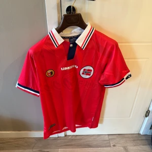 Norge fotbollströja Umbro röd - Röd Norge fotbollströja från Umbro med klassisk vit krage och blå detaljer. Broderad norsk flagga och förbundsmärke på bröstet, samt texten 'NORGE' på ärmen. Tillverkad i lätt och ventilerande polyester, perfekt för match eller träning. Helt ny