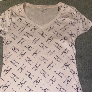 Ljusrosa t-shirt Tommy Hilfiger - Ljusrosa t-shirt från Tommy Hilfiger med korta ärmar och v-ringning. T-shirten har ett all-over tryck med Tommy Hilfiger-loggan i marinblått och rött. Skön passform och tillverkad i mjuk bomull., Aldrig använt. 