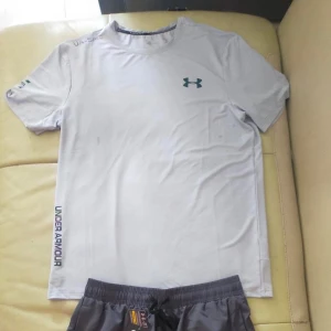 Under Armour reflective t shirt short set - Säljer ett snyggt grå sätt från Under Armour med logga på bröstet och tryck på ärmen, loggan på både shortsen och t shirten har reflex med regnbågsfärg. Shorts och T-shirten har rund hals, korta ärmar och är gjord i ett lätt och funktionellt material som passar perfekt till träning. Enkel och stilren design med sportig känsla. Priset är ej hugget i sten, finns i storlek L och M, passar även S och XL, (mitt tips är att ta en mindre storlek för dig som vill använda det som en compression gymshirt