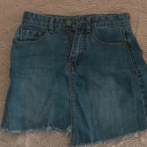 Blå jeanskjol med rå kant - Snygg blå jeanskjol med klassisk femficksmodell och rå, fransig kant nertill. Kjolen har bälteshällor, knapp och dragkedja framtill samt bakfickor. Perfekt för en chill och trendig look.