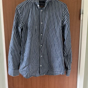 Randig blå skjorta från Riley - Snygg blå skjorta från Riley med vita vertikala ränder och slim fit-passform. Skjortan har långa ärmar, klassisk krage och knäppning framtill. Perfekt för dig som gillar stilrena och fräscha plagg med lite edge.