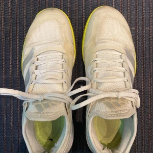 Adidas Adizero vita sneakers - Snygga Adidas Adizero sneakers i vitt med ljusgula detaljer på sulan. Skorna har klassisk snörning, mesh- och syntetmaterial för bra andningsförmåga och komfort. Perfekta för dig som gillar sportig stil och vill sticka ut med en fräsch färgkombination.