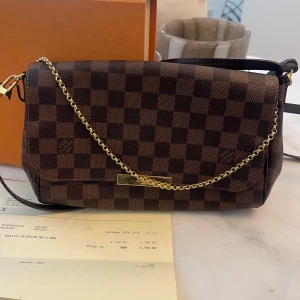 Louis Vuitton axelväska Damier Ebene - Säljer en snygg axelväska från Louis Vuitton i klassiska Damier Ebene-mönstret. Väskan är brun och mörkbrun med guldfärgad kedja och detaljer. Tillverkad i tåligt skinn och har en stilren, rektangulär form. Perfekt för dig som gillar lyxiga accessoarer.