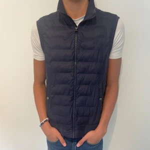 Mörkblå dunväst från Ralph Lauren - Snygg mörkblå dunväst från Ralph Lauren med quiltad framsida och dragkedja hela vägen. Västen har två fickor med knappstängning och diskret logga på bröstet. Perfekt att slänga över en hoodie eller t-shirt för en clean look. Nypris 2500kr skick 8/10 hör av er vid eventuella frågor 