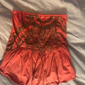 Korallröd tubtopp från Guess - Snygg korallröd tubtopp från Guess med coolt grafiskt mönster i svart och små nitar framtill. Toppen är axelbandslös och har lätt rynkade detaljer nedtill för en schysst siluett. Perfekt för dig som vill sticka ut med färg och detaljer.