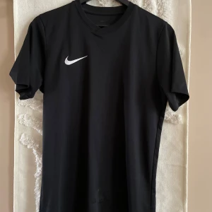 Svart Nike Dri-FIT fotbollströja - Svart fotbollströja från Nike med klassisk vit swoosh på bröstet. Tillverkad i Dri-FIT-material som andas och håller dig torr under matchen. Enkel och stilren design med rund hals och korta ärmar. Perfekt för träning eller match.