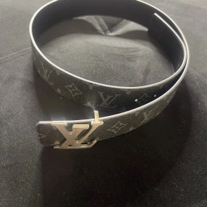 svart monogram bälte - Snyggt svart bälte med klassiskt monogrammönster och stor logga i silver som spänne. Bältet är gjort i skinn och har en stilren, rak form. Perfekt för dig som vill ha en lyxig accessoar med ikonisk design. Helt nytt med box! Bara för 300kr!