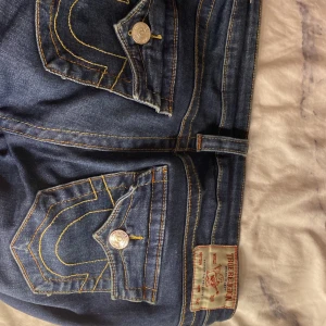 Low waist True Religion Joey jeans blå, stl 28 - Säljer ett par ikoniska True Religion Joey jeans i mörkblå denim med kontrastsömmar och stora fickor med lock och silverfärgade knappar. Klassisk bootcut-modell med låg midja och snygga detaljer. Perfekta för dig som gillar streetwear och vill sticka ut.