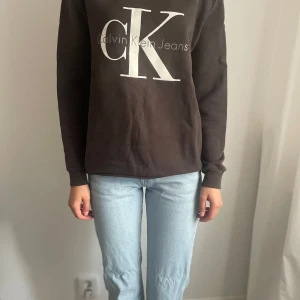 Brun sweatshirt från Calvin Klein Jeans - Mörkbrun sweatshirt från Calvin Klein Jeans med stor vit CK-logga framtill. Tröjan har rund hals, långa ärmar och är i mjuk bomullsblandning. Perfekt för en chill och stilren look.