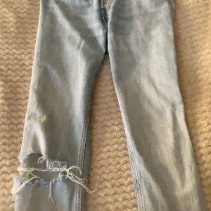 Ljusblå slitna jeans med hål - Säljer ett par ljusblå jeans med snyggt sliten look och stort hål med fransar på ena knät. Jeansen har rak passform och klassisk femficksdesign. Perfekta för dig som gillar en avslappnad och trendig stil.