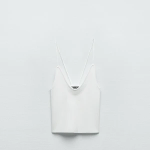 Vit stickad topp med smala axelband - Minimalistisk vit stickad topp med smala axelband och v-ringad hals. Toppen är croppad och har en stilren, enkel design som passar perfekt till jeans eller kjol. Perfekt för varma dagar eller lager på lager-stil.