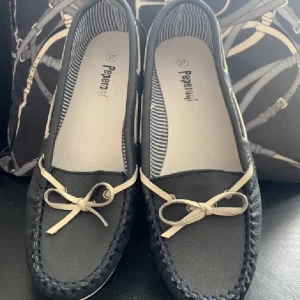 loafers med rosett från Peperoni - Stilrena  loafers från Peperoni med beige snördetalj och rosett framtill. Skorna har en platt vit sula och klassisk rund tå. Insidan är fodrad med randigt tyg för extra stilpoäng. Perfekta för en snygg och avslappnad look.