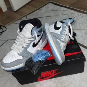 Nike Air Jordan 1 University Blue - Nike Air Jordan 1 High i färgen University Blue med vit och svart kontrast. Ovandel i vitt skinn och blå mocka, svart swoosh och klassisk Air Jordan-logga på sidan. Kommer med extra skosnören. Perfekta för dig som vill sticka ut med en ikonisk sneaker.