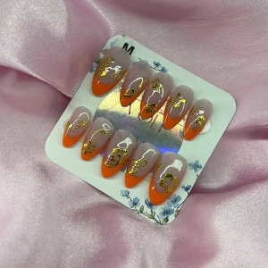Clementin (M);Medium Almond - Snygga gelé press-on naglar i medium mandelform med orangea french tippar och detaljerade guldfärgade blommor och mönster. Naglarna har en mjuk nude bas och kommer i storlek  M: 16 mm, 12 mm, 12,5 mm, 11,5 mm, 10 mm . Perfekt för dig som vill ha en trendig och unik look.
