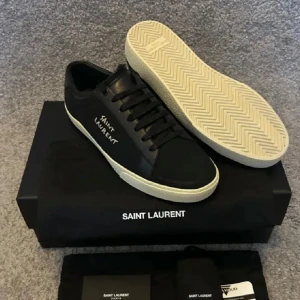 Svarta sneakers från Saint Laurent(Nya!) - Svarta sneakers från Saint Laurent med vit logga broderad på sidan. Skorna har svart snörning, rund tå och vit platt sula. Tillverkade i canvas och skinn för en clean och stilren look. Perfekta för dig som gillar minimalistisk design med lyxig känsla.
