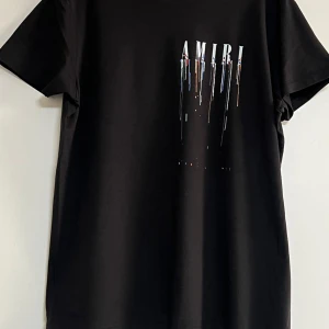 Svart Amiri t-shirt med ängeltryck - Svart t-shirt från Amiri med coolt tryck framtill där loggan droppar i olika färger och stor vit ängelgrafik på ryggen. Klassisk passform och korta ärmar, tillverkad i mjuk bomull. Perfekt statement-plagg för dig som gillar streetwear vibes.