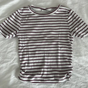 Randig t-shirt från Kappahl - Vit ribbad t-shirt från Kappahl med svarta och rosa horisontella ränder. Klassisk rund halsringning och korta ärmar. Skön och stretchig passform som funkar till det mesta. Perfekt för dig som gillar en enkel men ändå snygg stil.