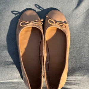 Beige ballerinaskor med rosett - Snygga beige ballerinaskor med rund tå och söt rosettdetalj framtill. Skorna har en klassisk, enkel design och är gjorda i ett glansigt syntetmaterial som ger en stilren look. Perfekta att matcha med både jeans och klänning.