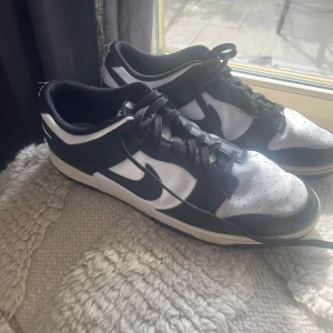 Nike Dunk Low svart/vit sneakers - Snygga Nike Dunk Low sneakers i svart och vitt med klassisk Swoosh-logga på sidan. Skorna har snörning, perforerad tå och låg profil. Perfekta för dig som gillar streetstyle och vill ha en ikonisk look. Tillverkade i syntet och skinn för en clean känsla.