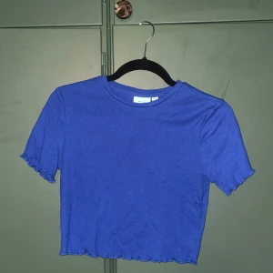 Blå croppad topp från Vila - En blå croppad topp från Vila med korta ärmar och vågiga kanter vid ärmslut och nederkant. T-shirten har en enkel, stilren design och är gjord i mjuk bomull. 