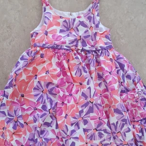 Klänning - Söt klänning med färgglatt blommönster i rosa, lila och vitt. Ärmlös modell med rund halsringning och lätt utställd kjol. Perfekt för varma dagar och ger en lekfull vibe.