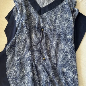 Blå mönstrad klänning Cover-up - Supersnygg blå klänning från Panos Emporio med vitt blommigt mönster och snörning i midjan. Lätt och luftig modell, perfekt för varma dagar. Klänningen har v-ringning och är tillverkad i ett mjukt material som känns skönt mot huden.