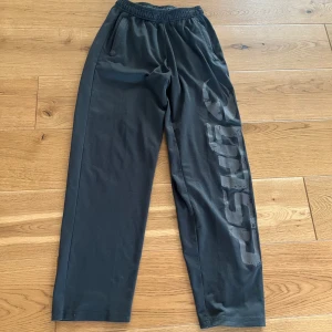 Svarta mjukisbyxor från GASP - Svarta mjukisbyxor från GASP med elastisk midja och stora fickor. Snyggt tryck längs ena benet och tillverkade i ett mjukt, syntetiskt material som passar perfekt för chill eller träning. Raka ben och avslappnad passform.