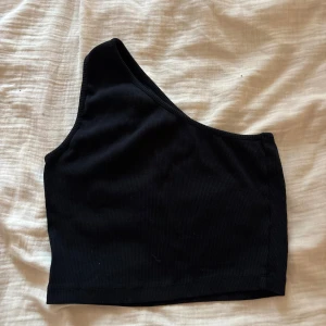 Svart one shoulder topp Even&Odd - Säljer en svart ribbad one shoulder topp från Even&Odd. Toppen är croppad och har en asymmetrisk design med en axel. Perfekt för dig som gillar stilrena och trendiga plagg. Materialet är mjukt och stretchigt för en bekväm passform.