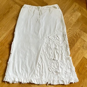Vit distressed långkjol - Vit lågmidjad långkjol med coola fransiga och råa detaljer längs nederkanten och på ena sidan. Kjolen har en rak passform och är tillverkad i ett mjukt bomullstyg. Liten fläck på baksidan(se bild). 
