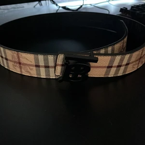 Burberry rutig bälte med B-spänne - Snyggt bälte från Burberry med klassiskt rutigt mönster i beige, svart, vitt och rött. Bältet har ett stilrent svart B-spänne framtill och är tillverkat i skinn med canvas-yttersida. Perfekt accessoar för att lyfta din outfit.