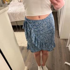 Blå blommig omlottkjol från SHEIN - Supersöt blå kortkjol från SHEIN. Kjolen har omlottmodell med knyt i sidan. Storleken står inte men sitter som en xs/s :)