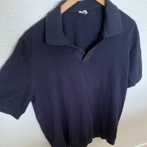 Svart pikétröja i bomull - En klassisk svart pikétröja med krage och korta ärmar. Tröjan är tillverkad i mjuk bomull och har en enkel, stilren design utan synliga loggor eller mönster. Perfekt för en clean och avslappnad look.