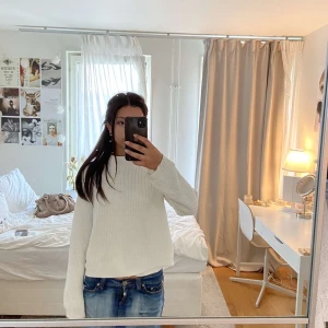 Vit stickad tröja med hög krage - Vit stickad tröja med högre krage och långärmad design. Tröjan har ett snyggt ribbat mönster på ärmar och kropp, vilket ger en cool struktur. Perfekt för lager-på-lager och passar till jeans eller kjol. Enkel och stilren look.