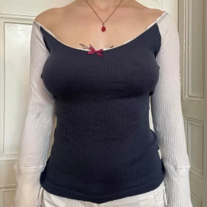 Mörkblå offshoulder topp med rosett - Superfin ribbad offshoulder topp i mörkblått med vita ärmar. Söt liten vinröd rosett framtill och spetskant vid urringningen. Långärmad och figurnära passform, perfekt för dig som gillar en unik och trendig look.