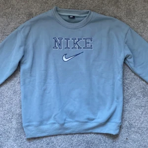 Ljusblå Nike sweatshirt i bomull - Ljusblå sweatshirt från Nike med klassisk logga och swoosh i vitt och blått framtill. Tröjan har rund hals, ribbade muddar och är tillverkad i mjuk bomullsmix. Perfekt för en chill och sportig look.