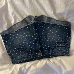 Jeansshorts - Jeansshorts med små vita hjärtan över hela tyget. Rå kant upptill för en cool look och detaljer som fickor och knappar framtill. Perfekt för dig som vill sticka ut med en kreativ och trendig stil.