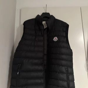 Svart dunväst från Moncler M - Säljer en svart dunväst från Moncler med klassisk quiltad design och hög krage. Västen har dragkedja framtill, två fickor med dragkedja och Moncler-logga på bröstet. Perfekt för lager-på-lager och snygg till streetwear.