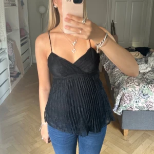 Svart spetsig linnetopp med tunna band - Supersöt svart linnetopp med tunna axelband och omlott framtill. Toppen är i spets med plisserad nederdel som ger en luftig och romantisk känsla. Perfekt att styla med jeans eller kjol för en trendig look.