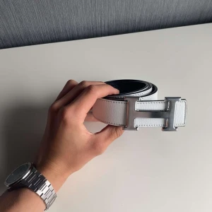 Vitt skärp med H-spänne - Snyggt vitt skärp med ikoniskt H-formad metallspänne i silver. Bältet är gjort i äkta skinn med en stilren och modern look. Perfekt accessoar för att lyfta din outfit med en exklusiv touch.