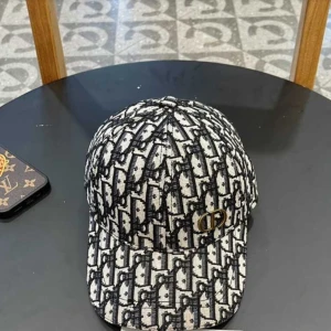 Dior keps med monogrammönster - Snygg keps från Dior med ikoniskt svart och vitt monogrammönster över hela kepsen. Kepsen har böjd skärm, justerbar rem baktill och en guldfärgad Dior-logga på sidan. Materialet är vävt tyg som ger en exklusiv känsla och streetstyle-vibe.