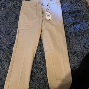 Beiga chinos Dublino slim fit Mango - Snygga beiga chinos från Mango i modellen Dublino slim fit. Byxorna har smart stil, regular längd och bälteshällor. Perfekta för en clean och stilren look. Tillverkade i ett mjukt bomullsmaterial.