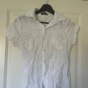 Vit kortärmad skjorta med knytband - Vit, broderad skjorta med korta ärmar, två bröstfickor och knappar framtill. Skjortan har ett knytband i midjan för snygg passform och ett diskret broderat mönster i tyget. Perfekt för en fräsch och somrig look. Inte hundra på storlek men skulle säga XS/S🤍