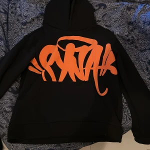 Svart hoodie SNTK med orange print - Svart hoodie från syna med stor, fet orange graffiti-inspirerad logga över bröstet. Klassisk huva och känguruficka framtill. Perfekt för dig som gillar streetwear och vill sticka ut med en cool design.